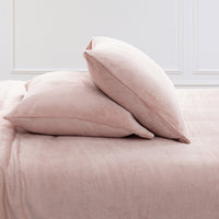 Two blush pink plush pillows resting on a matching pink bedspread against a white paneled wall.