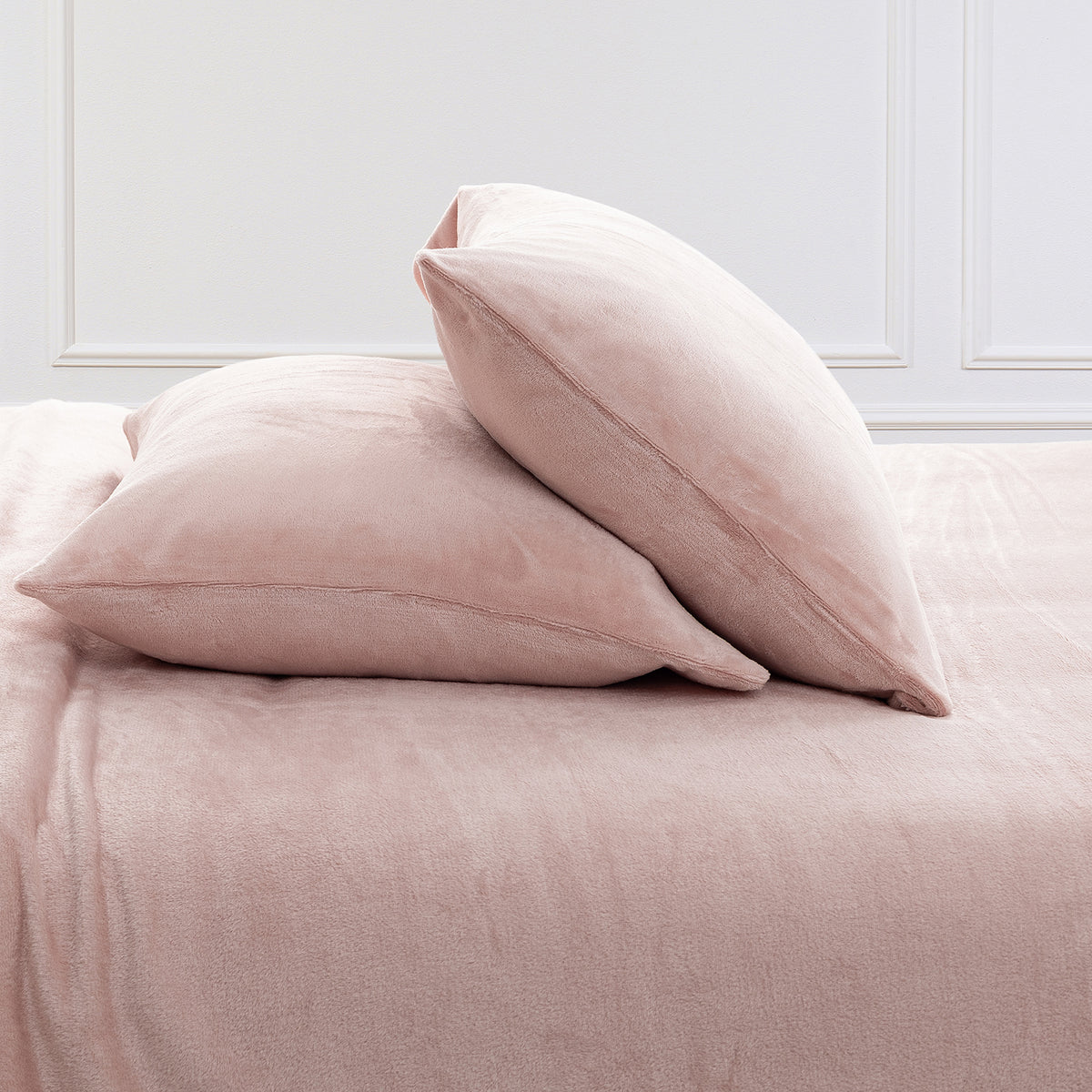 Two blush pink plush pillows resting on a matching pink bedspread against a white paneled wall.