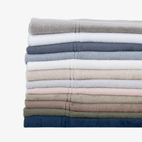 A stack of folded cotton towels in various muted colors, including gray, white, blue, beige, pink, brown, and navy, arranged vertically on a clean white background.