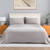 A neatly made bed in a styled bedroom setting, featuring a light gray throw blanket and two matching gray pillows on a white wooden headboard. The room has a gray wall, wooden nightstands, a white lamp, and a framed abstract print.