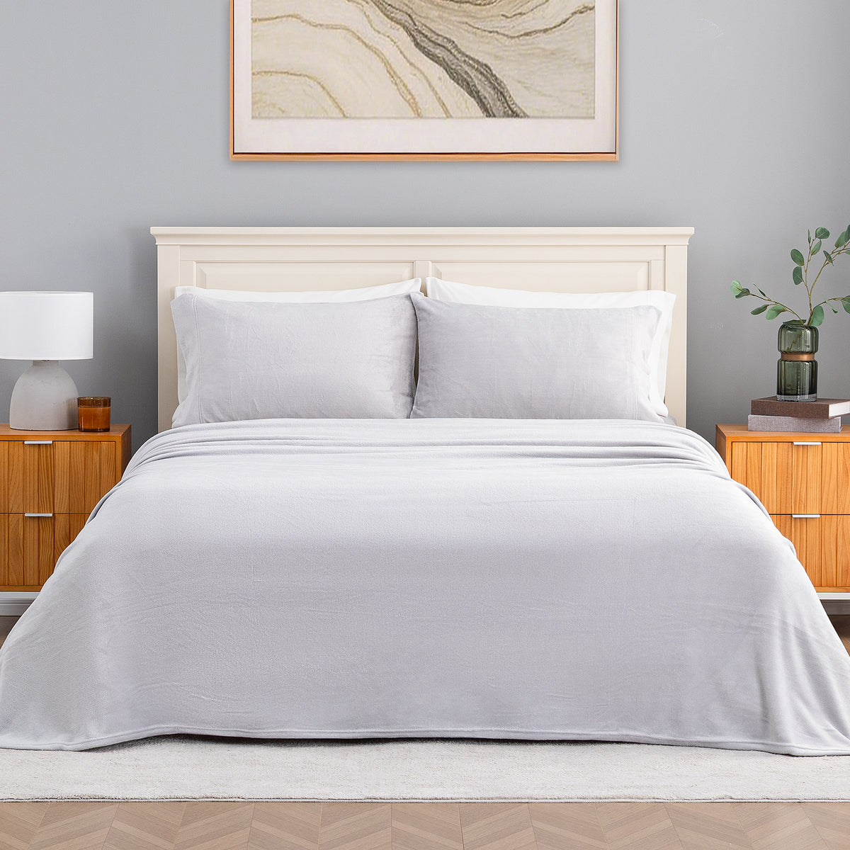 A neatly made bed with a light gray sheet and two matching pillows, styled in a bedroom with a cream headboard, wooden nightstands, a lamp, and framed wall art.
