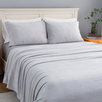 A neatly made bed with a light gray blanket and two matching pillow shams, set against a cream-colored wooden headboard in a softly lit bedroom.