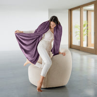 Woman sitting on a beige ottoman holding a purple throw blanket in a modern room.