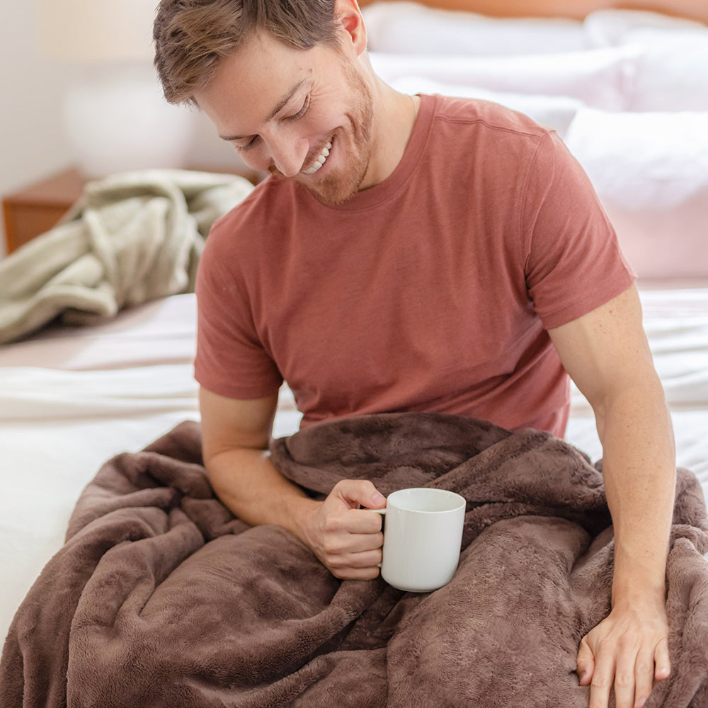 A man sitting on a bed with a brown PrimaLush Throw draped over his legs as he holds a mug.
