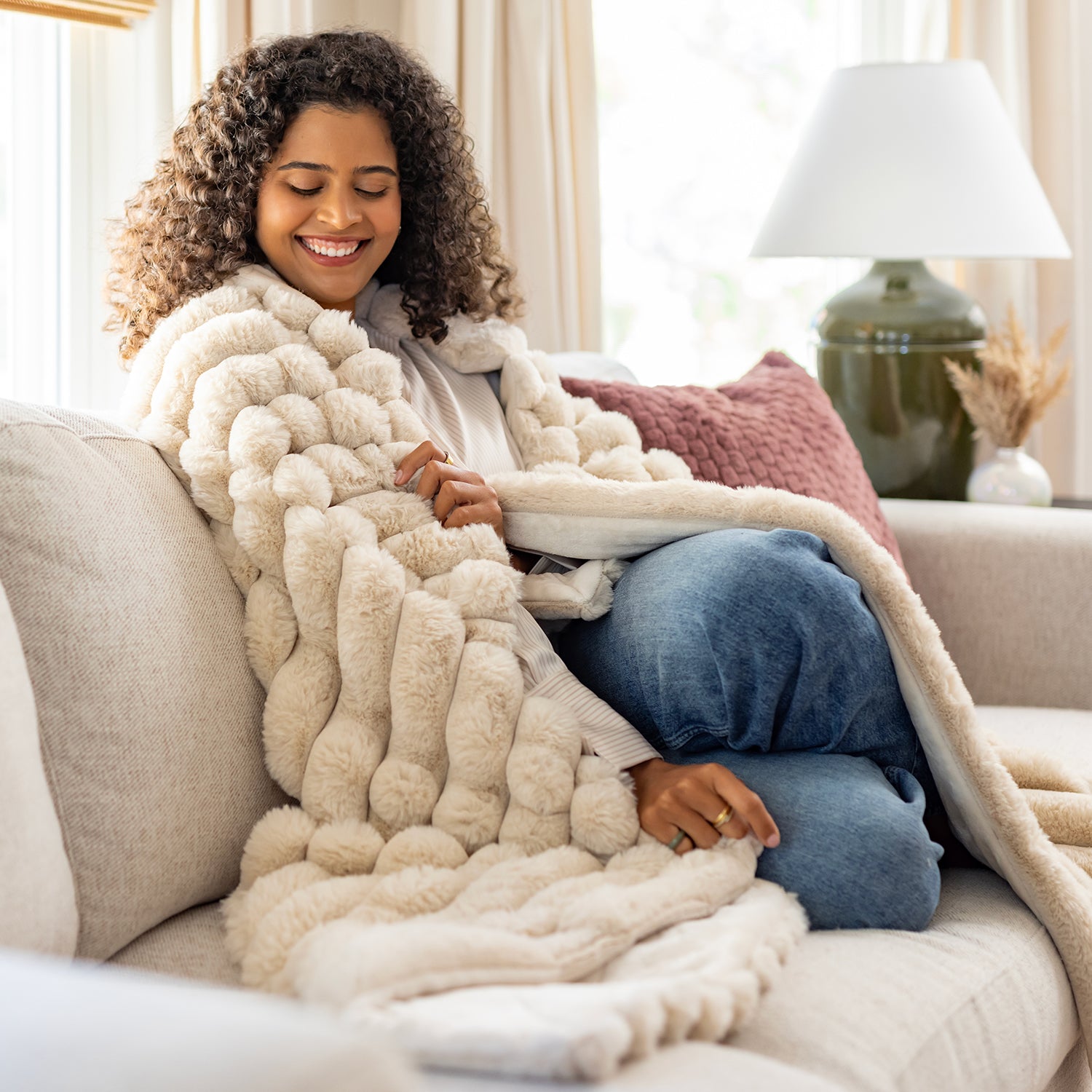 Woman sitting on a couch wrapped in a fluffy beige blanket, smiling.