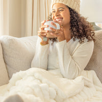 Woman sitting on a couch, wrapped in a blanket, holding a mug and smiling.