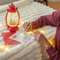 Person writing a letter next to a red lantern and cards on a soft surface with lights.