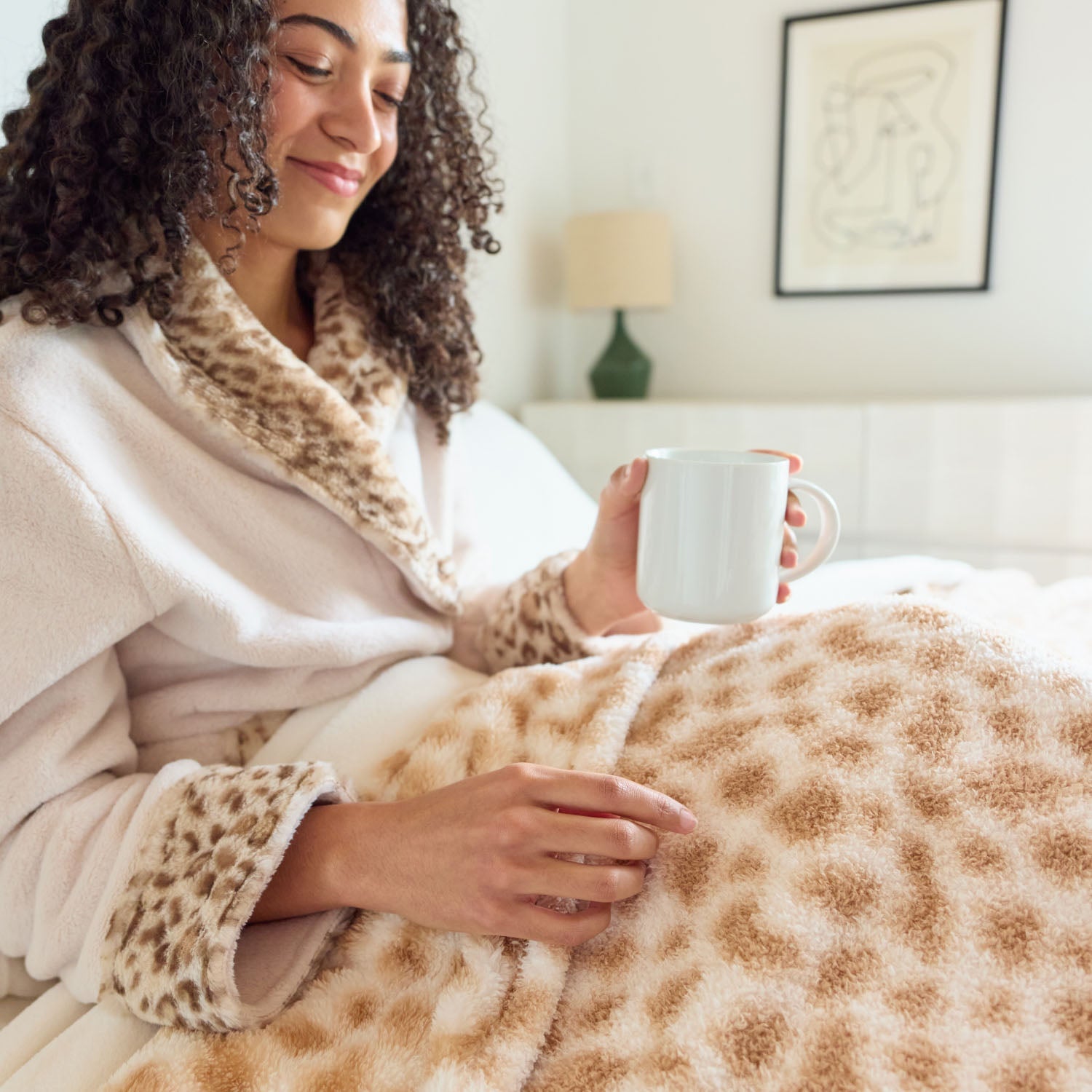 Woman in a cozy robe holding a mug, wrapped in a leopard print blanket in a home setting.