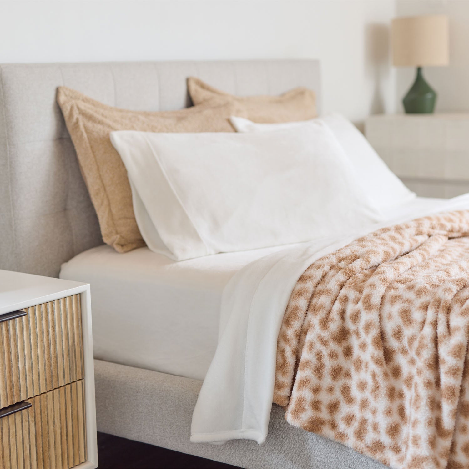 Neatly made bed with white pillows and a leopard print blanket next to a wooden nightstand.