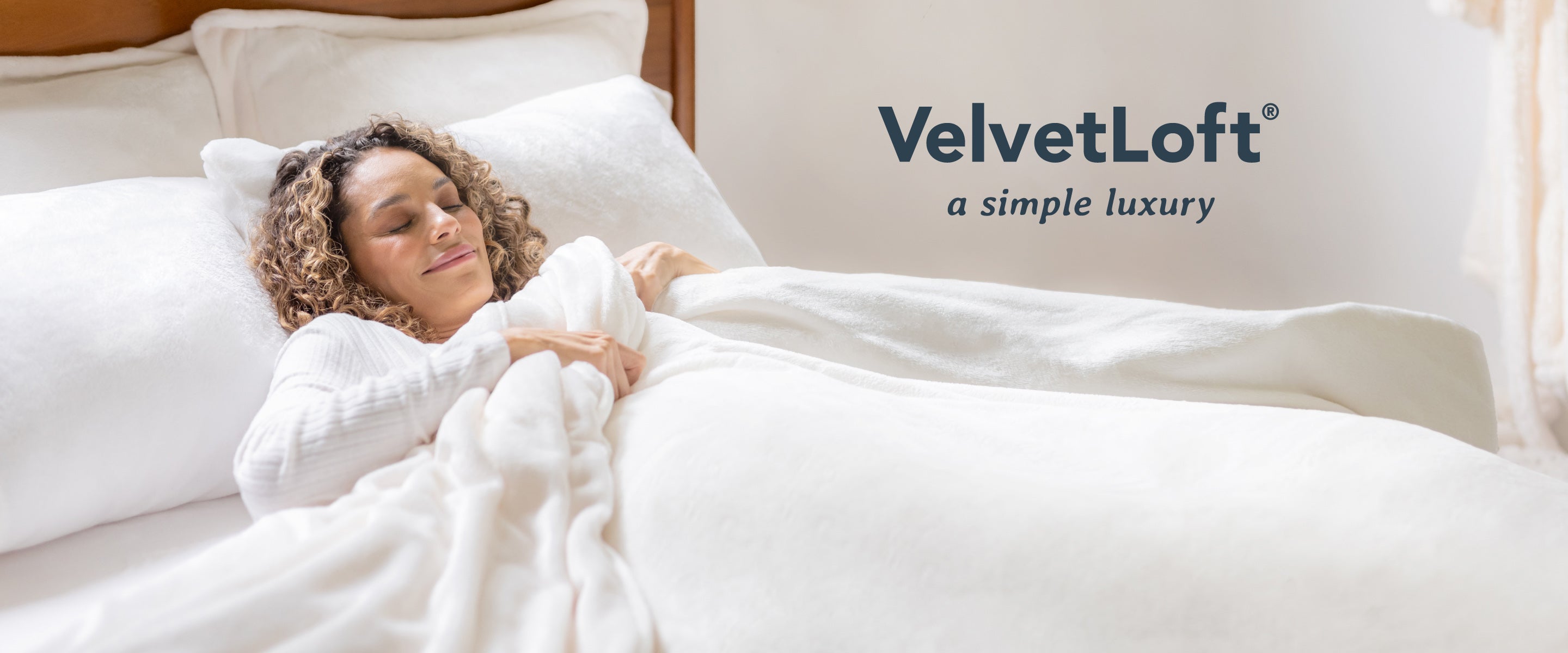 VelvetLoft® - a simple luxury. Image of a woman with curly hair tucked into an all white bed made with VelvetLoft® products. 